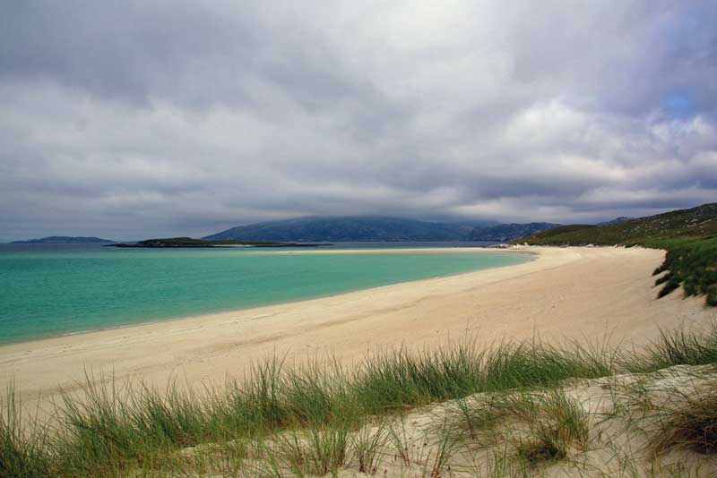 Under a cloudy sky the sand at Traigh Mheilein looks yellow. The water which is on the left hand side looks turquoise. In the foreground is some green beach grass