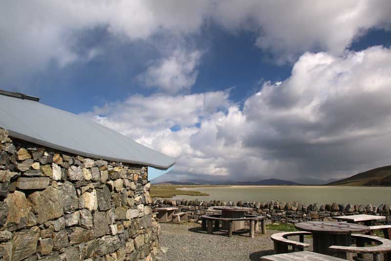 Temple cafe in Northton is built in stone. Outside are wooden tables which looks out over the sea. It is sunny but in the distance are dark clouds and it is raining