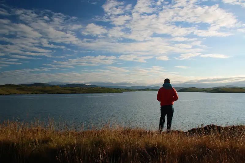 Man wearing an orange jacket standing looking out over a fjord in the evening with blue sky with several white clouds