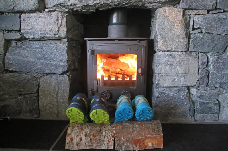 One pair of dark blue and green and one pair of light blue hiking shoes is drying in front a fire